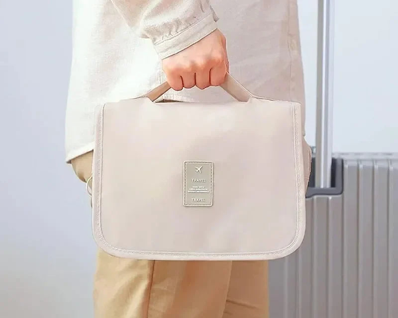 Trousse de toilette beige tenue par sa poignée à côté d'une valise
