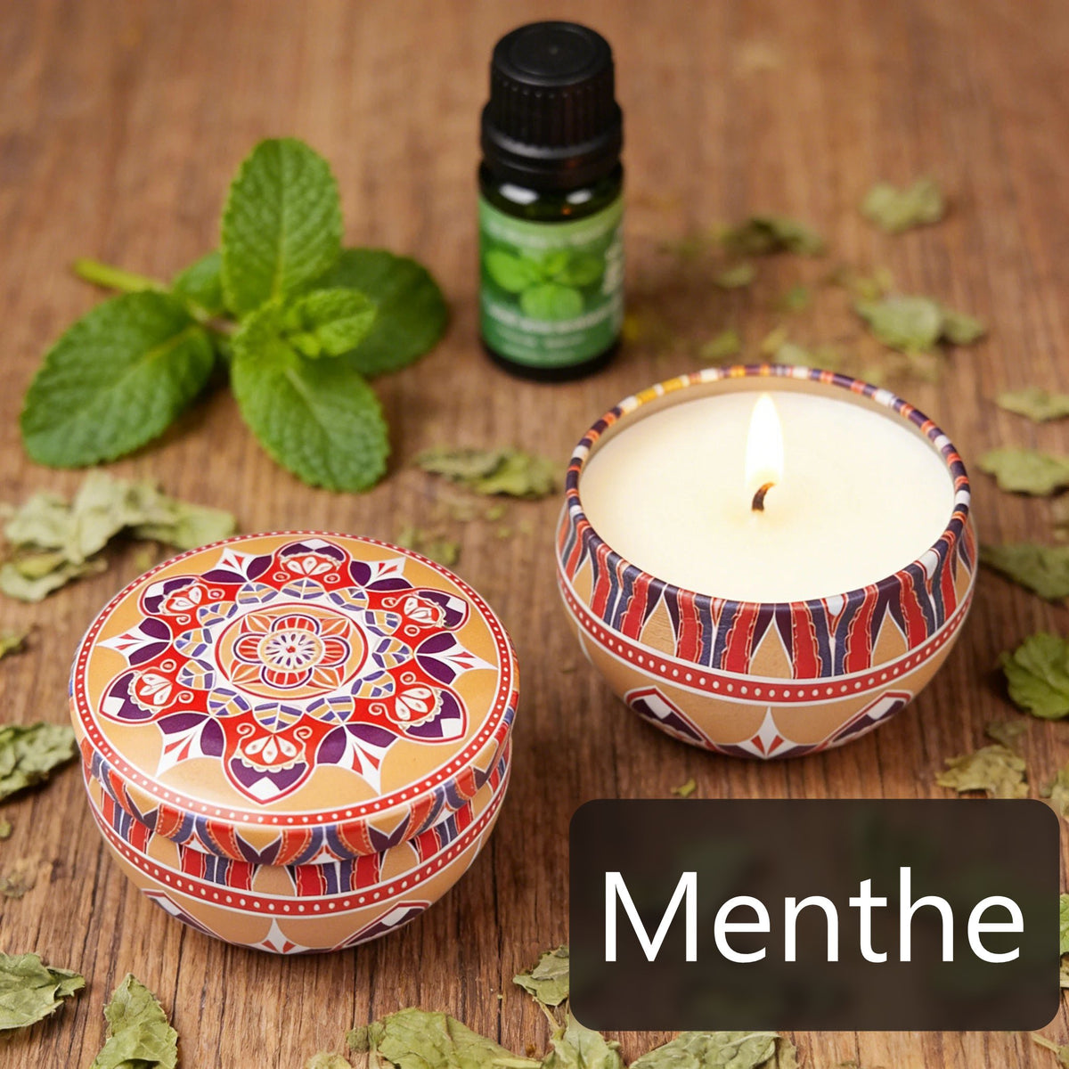 Deux bougies en boîtes métalliques décorées, feuilles de menthe et flacon d’huile sur une table en bois.